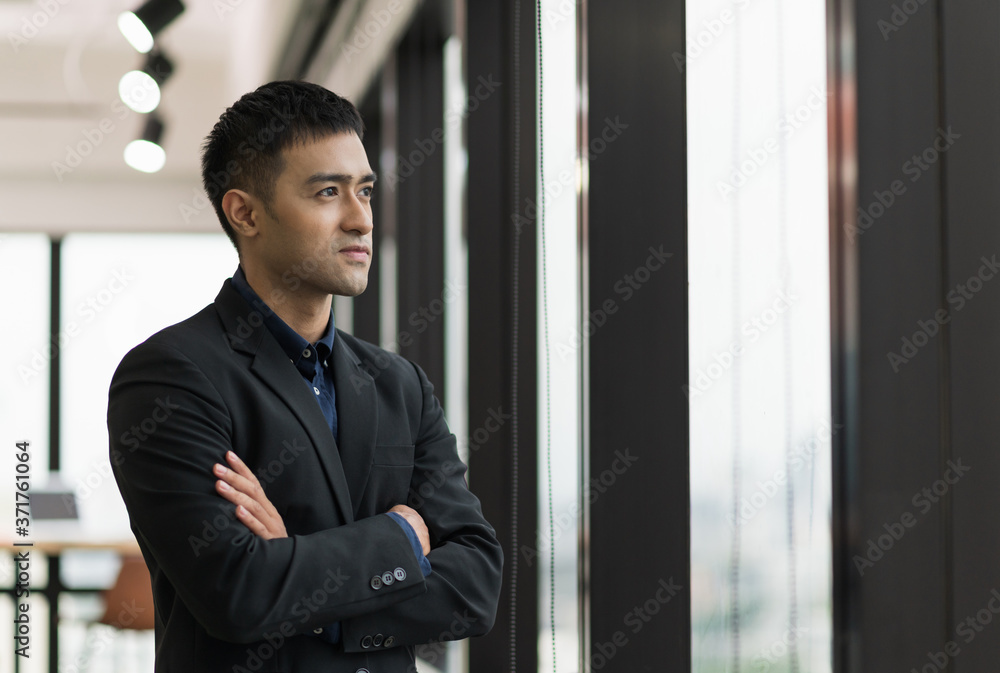 Young handsome asian businessman in suit standing near window with arm crossed in office. Young handsome asian businessman in suit standing near window with arm crossed in office.