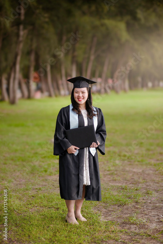 Happy and excited portrait of young Asian student with graduation hat in the outdoor.Smiling female Chinese wearing mortarboard and gown while holding certificate file with two hands.