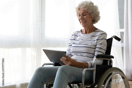 Billede på lærred Happy old mature disabled woman sitting in invalid carriage, using computer tablet, enjoying communicating with friends online, watching funny movie or TV series indoors, handicapped people lifestyle
