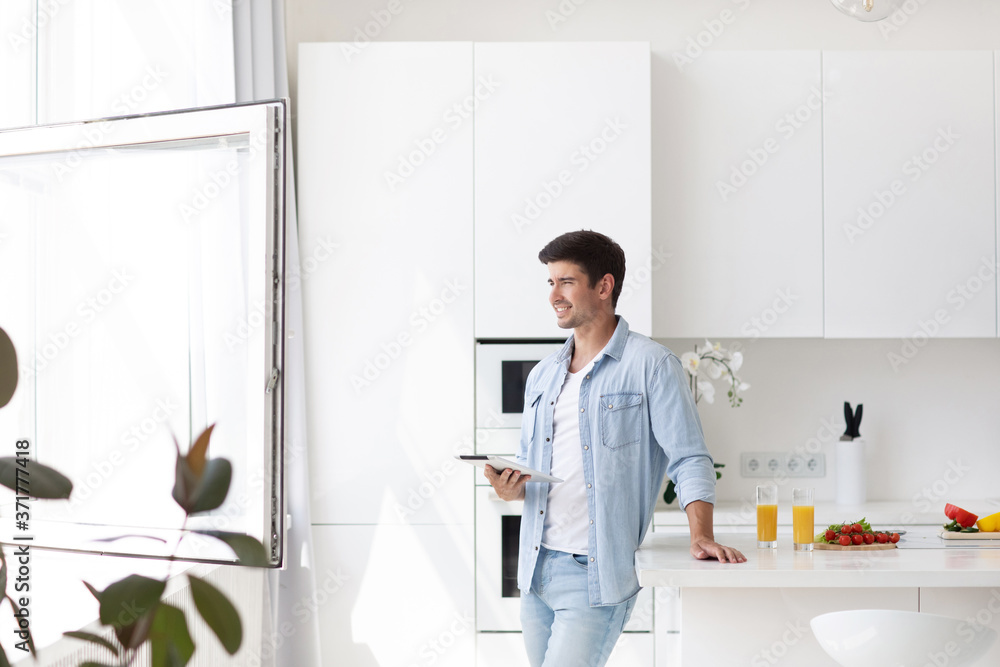 Young man standing on domestic kitchen holds digital tablet searching recipes for cook vegetable salad healthy food.