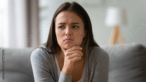Head shot close up unhappy young brunette frowning woman worrying about difficult decision, sitting on couch indoors. Frustrated millennial lady suffering from personal psychological problem.