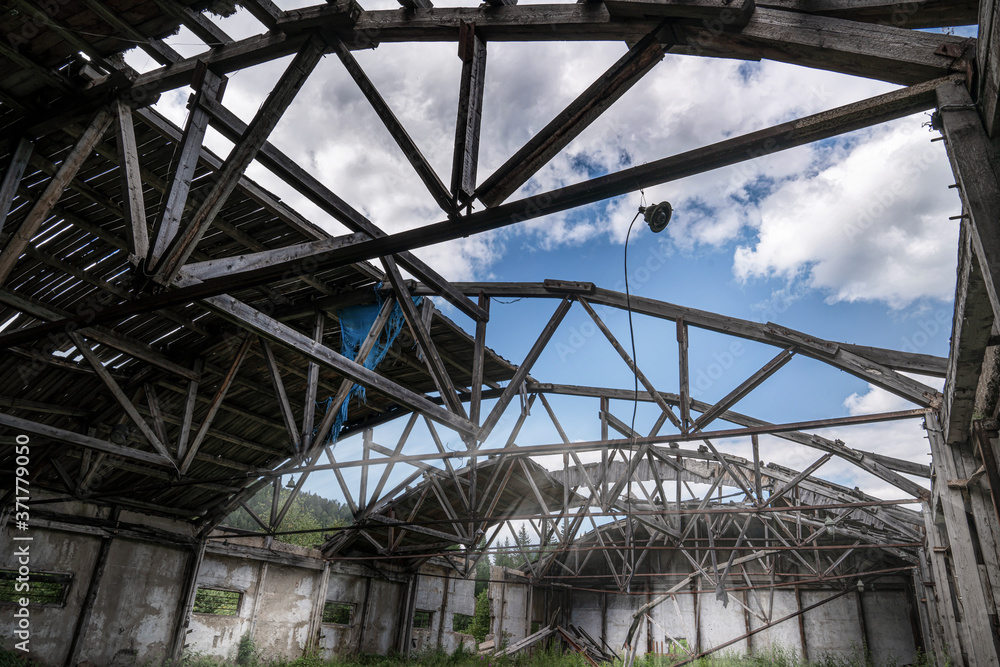 an old abandoned warehouse hangar with a ruined leaky roof and wooden floors, overgrown with grass inside. you can see the cloudy sky through the roof