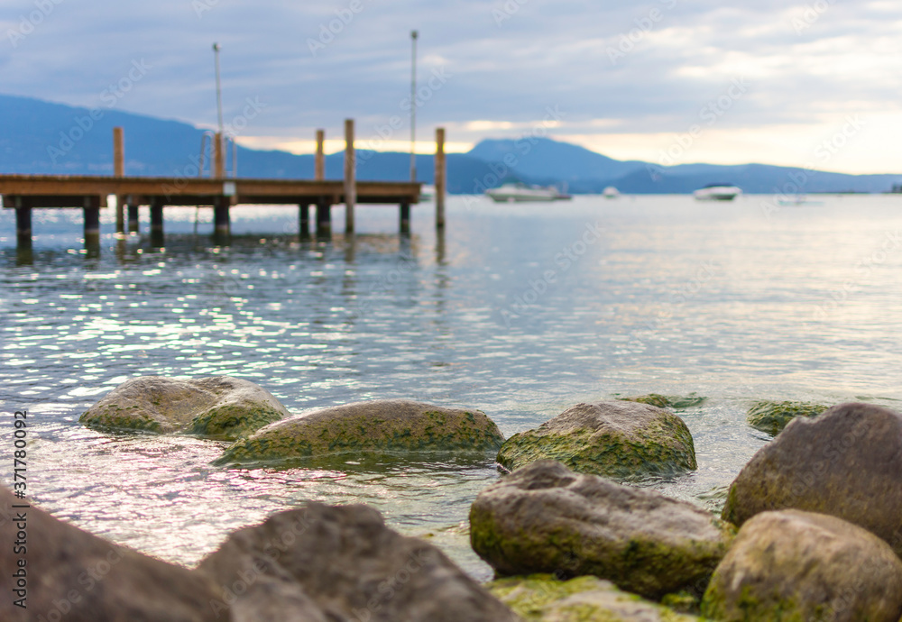 In the background a boardwalk leads into Lake Garda. In the foreground are large pebbles and stones overgrown with algae. The sky is cloudy. In the background a boardwalk leads into Lake Garda. In the foreground are large pebbles and stones overgrown with algae. The sky is cloudy.