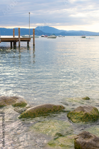 In the background a boardwalk leads into Lake Garda. In the foreground are large pebbles and stones overgrown with algae. The sky is cloudy.