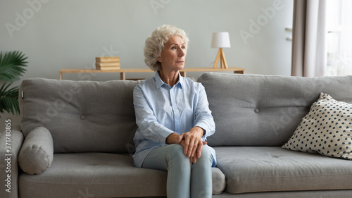 Thoughtful mature elderly hoary woman sitting on comfortable couch with folded hands on knees, suffering from mental disorder at home. Pensive older senior grandmother feeling loneliness indoors. © fizkes