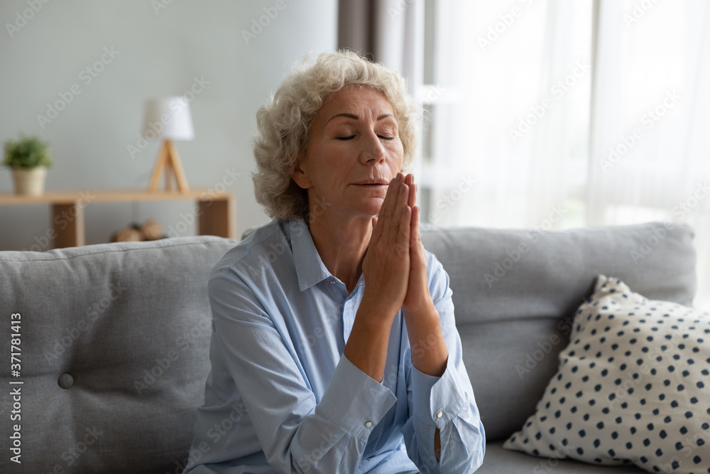 Peaceful middle aged woman sitting on sofa with folded in amen hands, feeling grateful indoors. Faithful senior mature praying God, asking for help, waiting for miracle or begging for forgiveness. Peaceful middle aged woman sitting on sofa with folded in amen hands, feeling grateful indoors. Faithful senior mature praying God, asking for help, waiting for miracle or begging for forgiveness.
