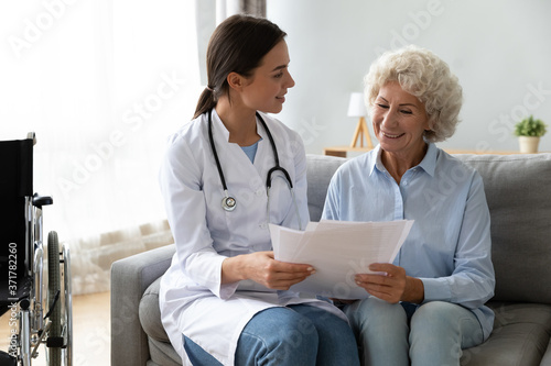 Happy young skilled female general practitioner in white medic uniform visiting disabled smiling middle aged senior patient, showing health analysis on paper, satisfied with treatment results.