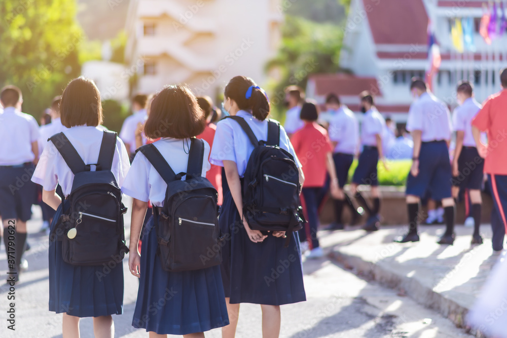 The back view of Asian high school students in white uniform wearing masks are walking towards preparing to respect the national flag in the morning amidst the beautiful nature surrounding the school. The back view of Asian high school students in white uniform wearing masks are walking towards preparing to respect the national flag in the morning amidst the beautiful nature surrounding the school.