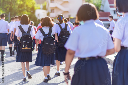The back view of Asian high school students in white uniform wearing masks are walking towards preparing to respect the national flag in the morning amidst the beautiful nature surrounding the school.