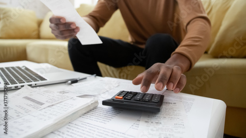 Black small business owner making calculations at home. Close up of african american man checking bills, counting expences, examining credit balance or planning household sitting on sofa in living photo