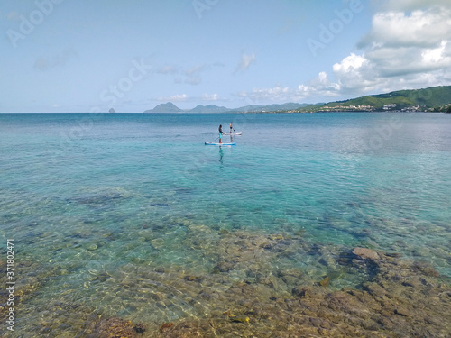 Idyllic landscape of the calm Caribbean Sea with a couple practicing paddle surfing under a tropical blue sky. Tropical and Caribbean landscape background of the French West Indies.
