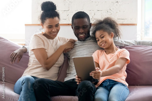 Mommy, daddy, level up. Cute happy african school age girl resting with parents on cozy sofa at living room holding digital pad, showing skills in playing online game, watching funny video or cartoon