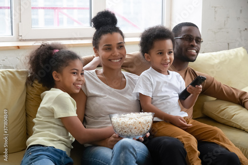 Family movie. Happy african couple with little daughter and son sitting on sofa hugging and looking on tv screen, smiling black family with children relax at home together watching funny show or film