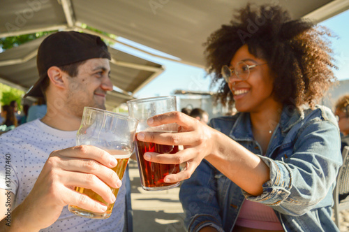 Young beautiful happy Caucasian and Afro couple of friends toasting with glasses of beers outside. Smiling while cheering together. Concept about people, lifestyle and holiday. 
