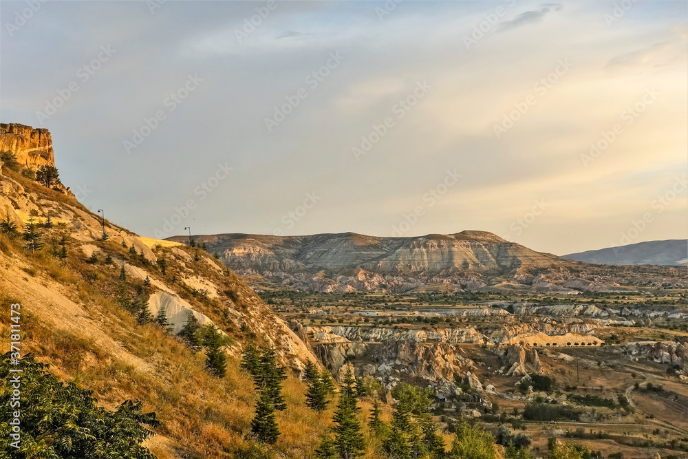 Cappadocia landscape before sunset. Mountains of unusual shape are painted in golden tones. Peaked rocks and trees are visible in the valley. Beautiful sky. Turkey.
