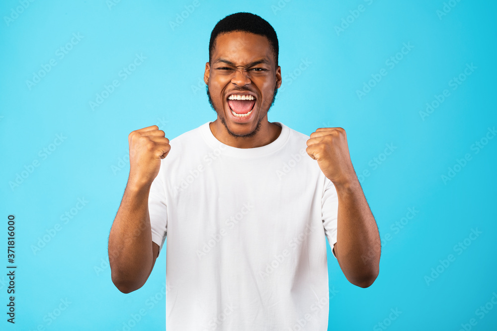 Fototapeta premium Emotional Black Guy Shouting Yes Shaking Fists On Blue Background