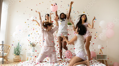 Pajama or bachelorette party celebration concept. Five slim active multiethnic women wearing comfy nightwear sexy glamor pyjamas hanging out jumping dancing on bed under falling multi colored confetti photo