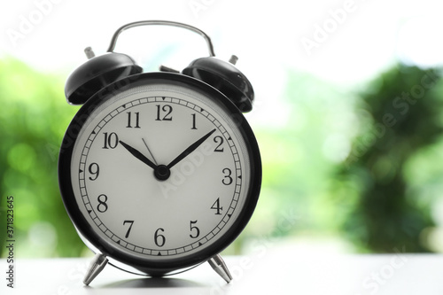 Modern black alarm clock on white table against blurred background. Space for text
