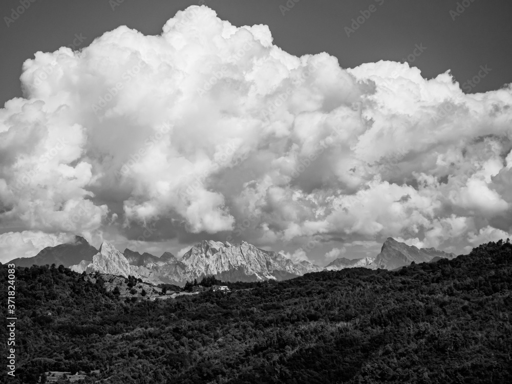 Black and white shot of rocky mountains on the background with clouds above.
In the foreground there are woods covered mountains with some visible houses Black and white shot of rocky mountains on the background with clouds above.
In the foreground there are woods covered mountains with some visible houses