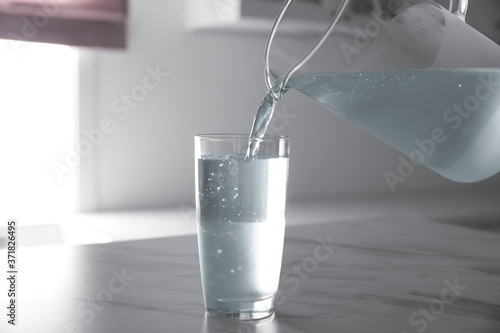 Pouring water from jug into glass on white marble table in kitchen
