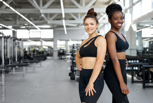 portrait of two diverse sportive girls in gym, young african and caucasian women in sportswear look at camera and smile, happy to lead healthy lifestyle