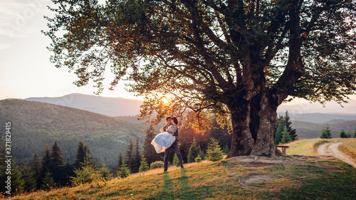 Fototapeta Naklejka Na Ścianę i Meble -  Loving newlyweds couple hugging in mountains at sunset. Groom holding bride on hands. Wedding in summer Carpathians.