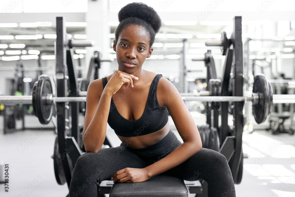 portrait of black woman taking a break after workout at gym. she sits next to barbells, dumbbells, posing at camera, wearing wportswear portrait of black woman taking a break after workout at gym. she sits next to barbells, dumbbells, posing at camera, wearing wportswear