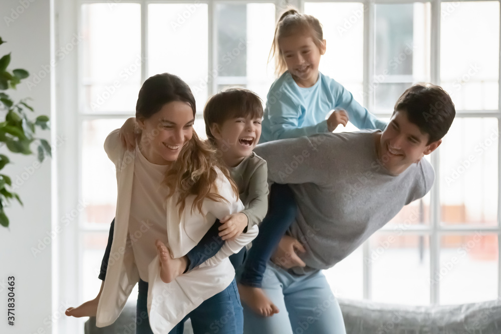 Playful young mixed race mother holding on back emotional little kid son while smiling father giving piggyback ride to happy preschool daughter, parents playing with energetic children at home. Playful young mixed race mother holding on back emotional little kid son while smiling father giving piggyback ride to happy preschool daughter, parents playing with energetic children at home.