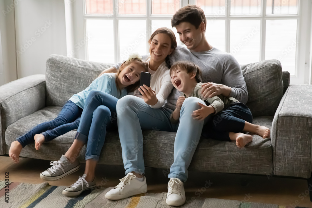 Smiling loving young couple parents showing funny video or watching childish cartoons on smartphone with laughing adorable kids siblings, enjoying stress free leisure time together on sofa at home. Smiling loving young couple parents showing funny video or watching childish cartoons on smartphone with laughing adorable kids siblings, enjoying stress free leisure time together on sofa at home.