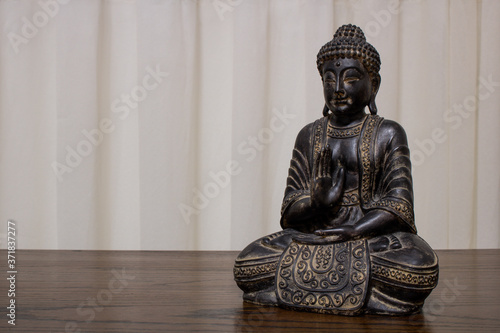 Dusty Buddha statue sitting in meditation on wooden table with soft textured beige curtains in the background. Tranquil and minimalist setting with warm tones and empty space for text