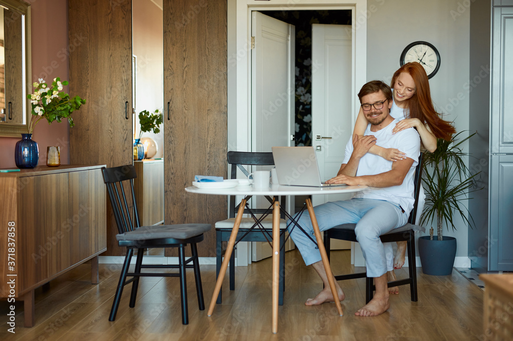 woman tries to cheer up her husband during his work on laptop, man sit with laptop, he is on freelance. redhead lady loves hes husband woman tries to cheer up her husband during his work on laptop, man sit with laptop, he is on freelance. redhead lady loves hes husband