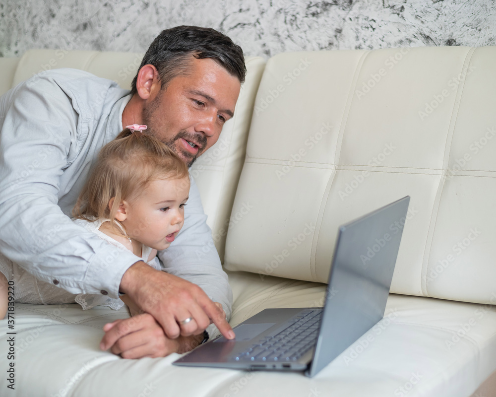 Little girl and caring father are watching cartoons on a laptop while lying on the couch at home. A man shows his little daughter photographs on a computer. Beloved daddy. Little girl and caring father are watching cartoons on a laptop while lying on the couch at home. A man shows his little daughter photographs on a computer. Beloved daddy.