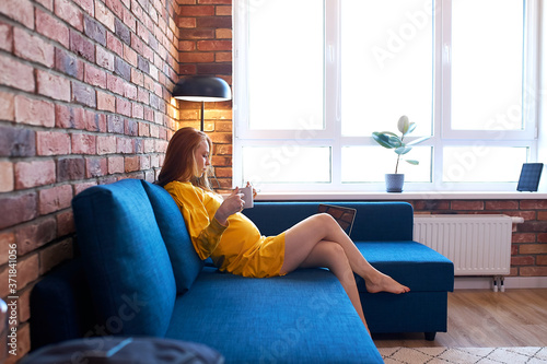 side view on attractive redhead pregnant woman drinking tea at home, woman in yellow dress sits on sofa, have rest