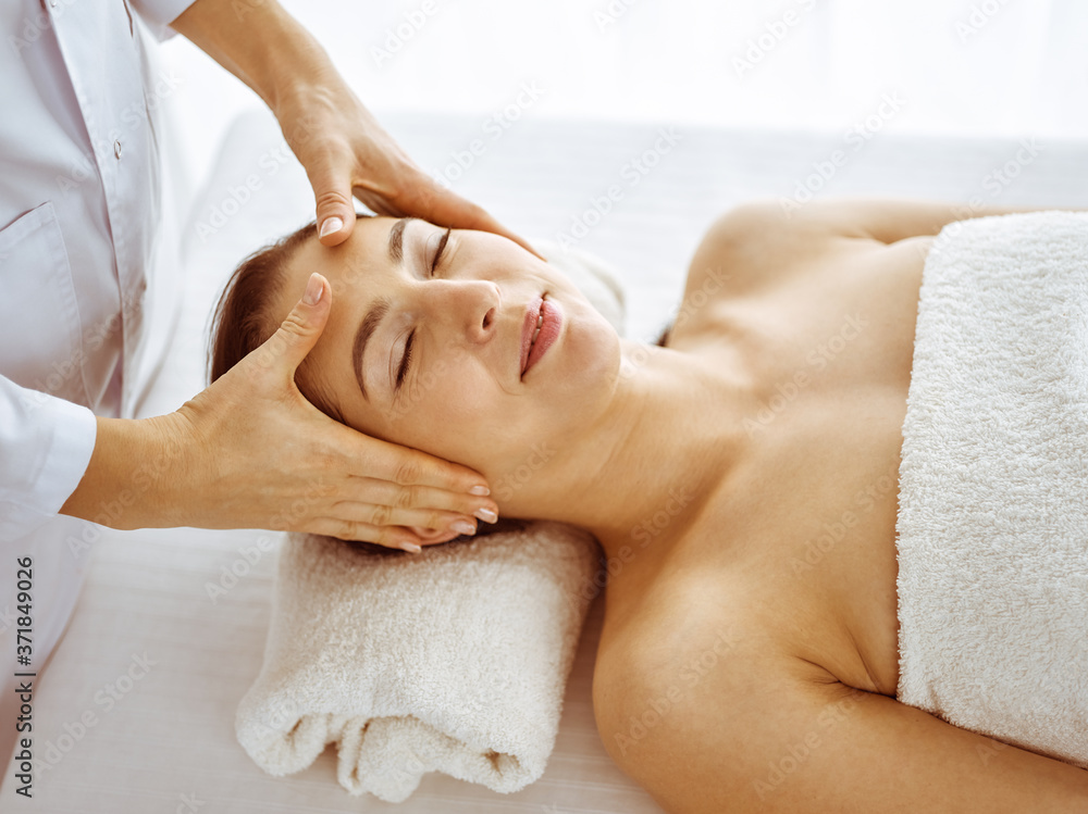 Fototapeta premium Beautiful brunette woman enjoying facial massage with closed eyes. Relaxing treatment in medicine and spa center concepts