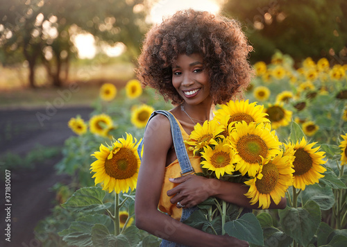 Happy young black woman walks in the sunflower field. Smiling dark-skinned girl with with a bouquet of sunflowers and curly hair.