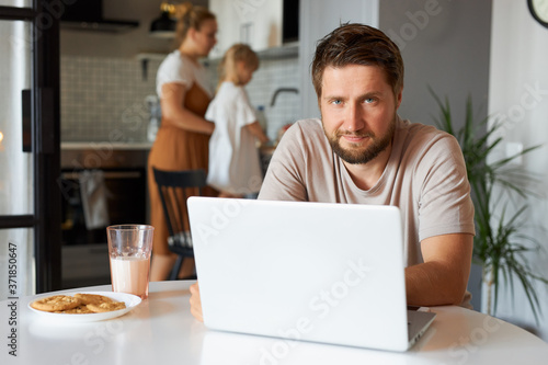 confident caucasian male work from home, sit in the kitchen with laptop, while his wife and daughter doing household chores in the background