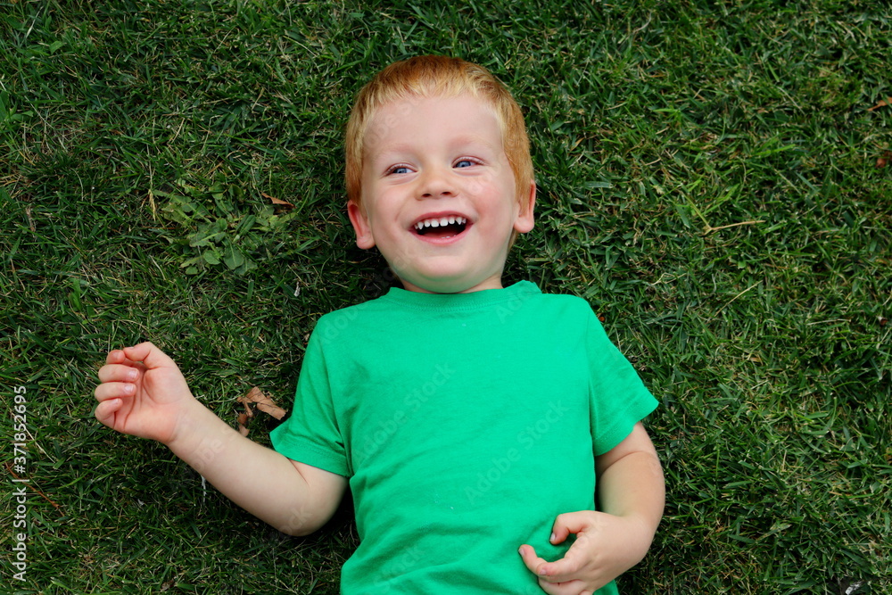 The blond boy lies on the green grass and smiles. Portrait of a three-year-old boy. Happy child. The blond boy lies on the green grass and smiles. Portrait of a three-year-old boy. Happy child.