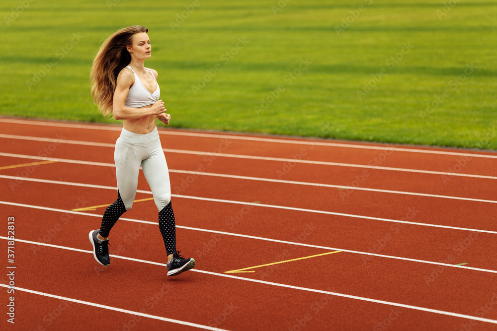 Obraz premium Athletic woman running at the stadium, working out, keep herself in shape, wearing sportswear, white top and pants, recommends doing sports, healthy lifestyle concept