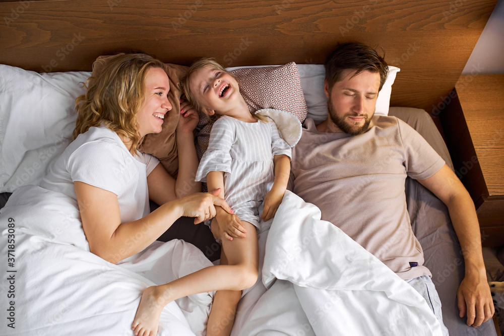 mother and daughter have fun on bed while father is sleeping, family on bed, weekends concept mother and daughter have fun on bed while father is sleeping, family on bed, weekends concept