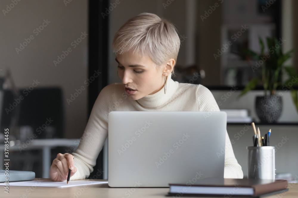 Front view focused young businesswoman sitting at table with computer, reading contract terms of conditions, checking paper report, analyzing marketing research results or editing document in office. Front view focused young businesswoman sitting at table with computer, reading contract terms of conditions, checking paper report, analyzing marketing research results or editing document in office.
