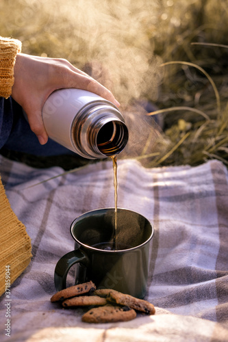 Women pouring hot tea coffee with steam  from thermos to cup standing on the cozy plaid - autumn picnic outdoor