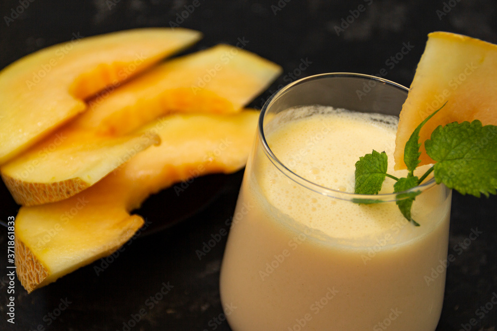 Melon milk shake on a black background. Ripe sliced melon is lying nearby. The smoothie glass is decorated with a sprig of mint and melon crumbs. Summer healthy food concept Melon milk shake on a black background. Ripe sliced melon is lying nearby. The smoothie glass is decorated with a sprig of mint and melon crumbs. Summer healthy food concept