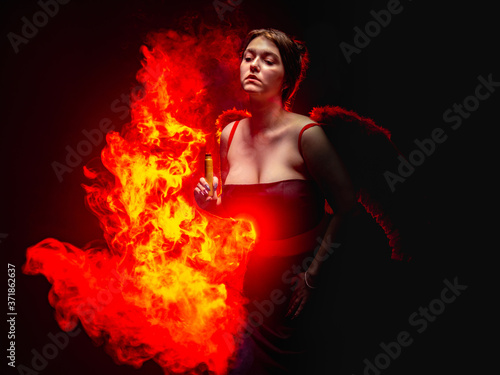 Devil. Girl with e-cig on the fire background. A woman in a devil costume. A young woman depicts a devil with a vape device. Red wings behind the dancer's back. Witch with fire. Devilish fire.