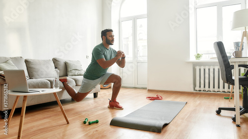 Get Fit At Home. Full length shot of young active man watching online video training on laptop, exercising, stretching during morning workout at home. Sport, healthy lifestyle photo