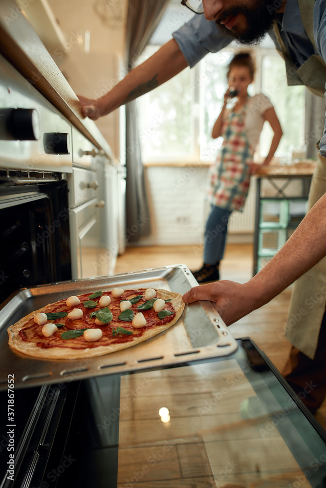 Professional cook making pizza at home. Man putting raw pizza in modern oven for baking. Woman standing in the background. Hobby, lifestyle