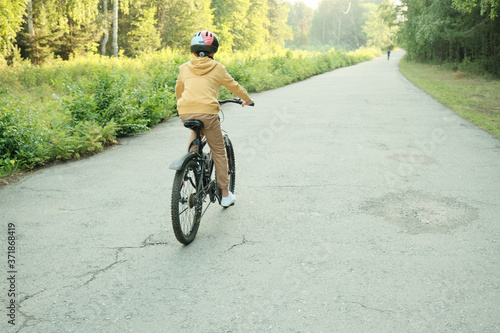 Rear view of youthful boy in casualwear and protective helmet riding bicycle