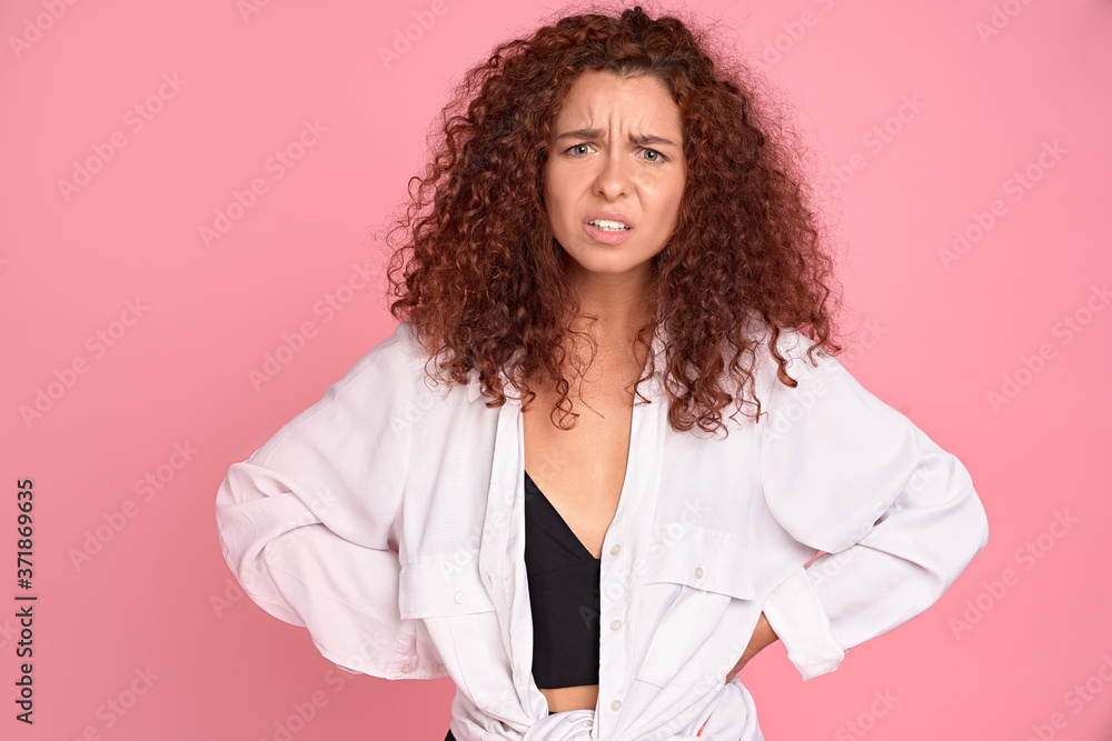 Young displeased redhead girl gesturing looking at camera. Confused girl arguing with a boyfriend. Pink background.