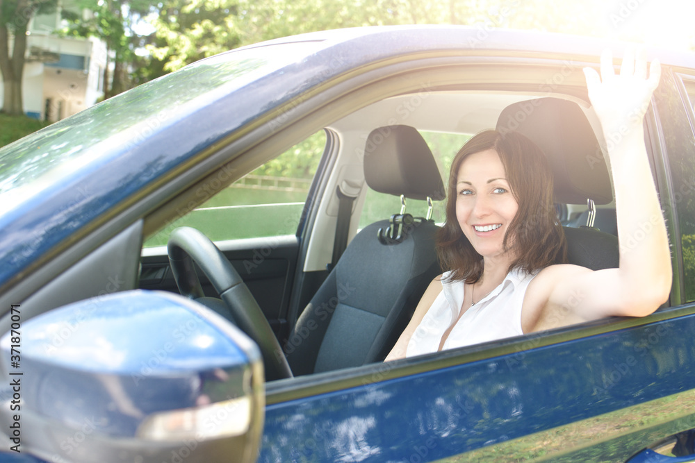 Pretty girl is waving hand, giving smile to someone. say hello, Beautiful happy adult woman sits in the car and waving his hand in greeting