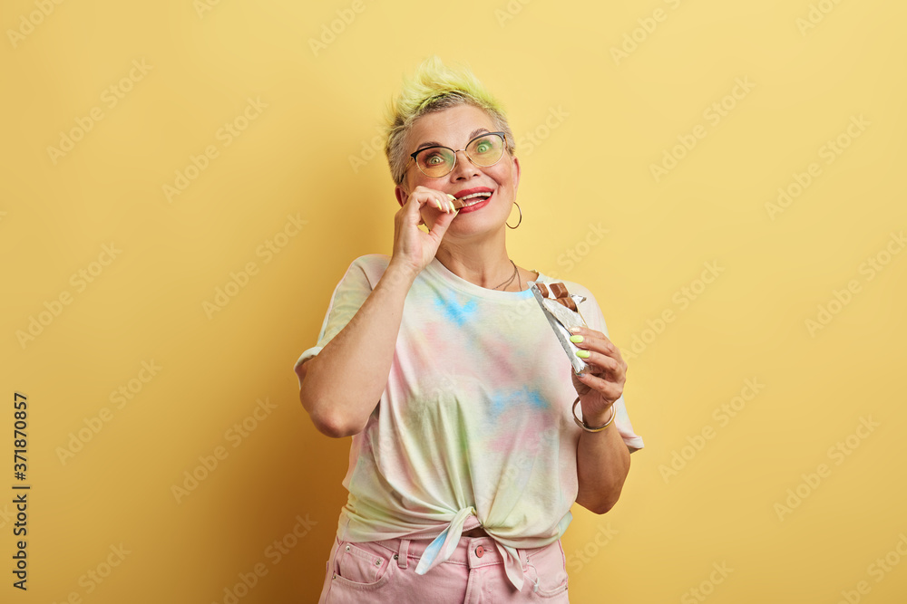 young glamour old woman enjoying eating chocolate isolated over yellow wall background, old woman doesn't care about her weights, favourite food, sweet tooth, lifestyle young glamour old woman enjoying eating chocolate isolated over yellow wall background, old woman doesn't care about her weights, favourite food, sweet tooth, lifestyle
