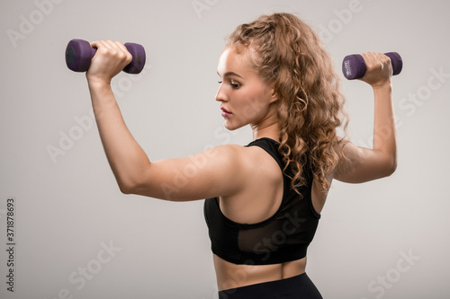 Back view of fit girl with long blond curly hair exercising with dumbbells